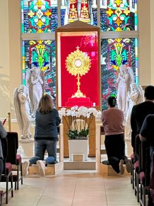 Monstrance with the Eucharist on an altar surrounded by angels and 2 women kneeling in prayer and adoration