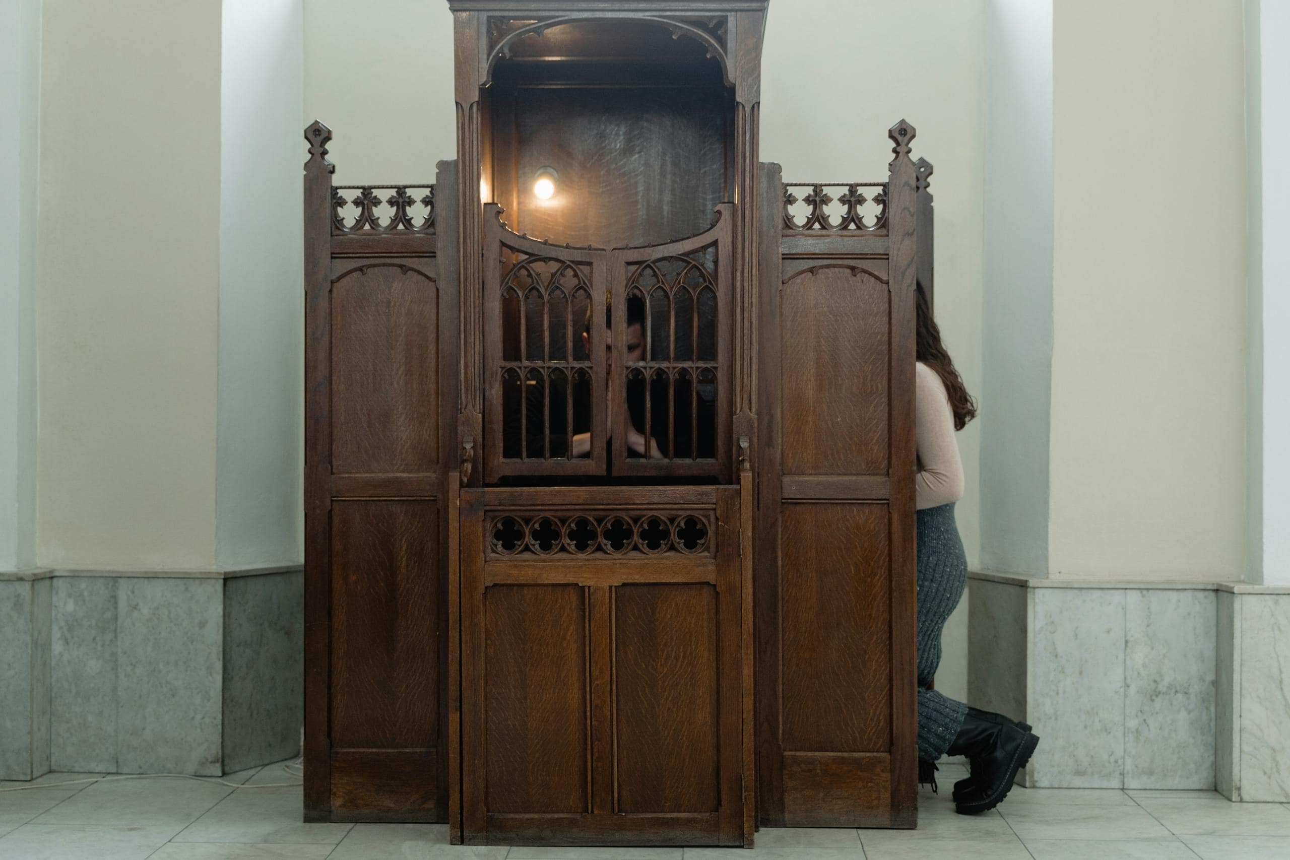 A wooden confessional booth in a church with a person sitting inside, partially visible.