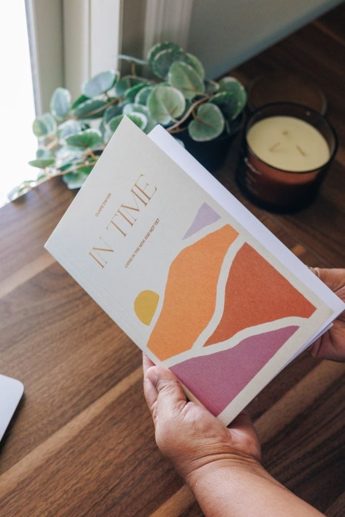 A woman holding a magazine titled "IN TIME" with abstract art on the cover, near a window with a pla.
