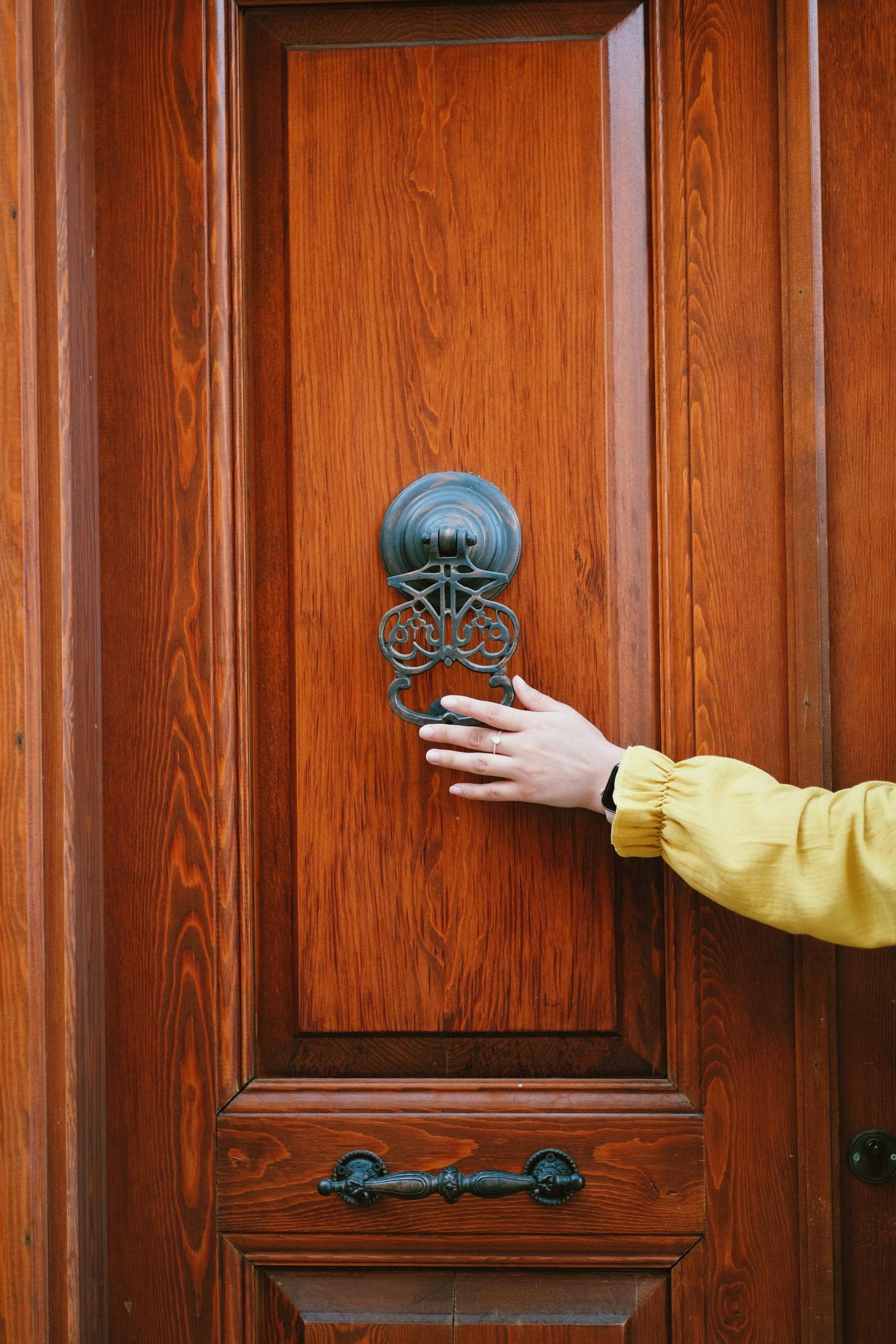 A person in a yellow sleeve reaching for a classic door knocker on a wooden door.